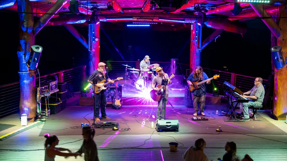 This exterior image shows musicians performing on an outdoor stage under colorful lights, with an enthusiastic audience dancing and socializing in the foreground. The setting captures the vibrant energy of a community concert or gathering, surrounded by the natural beauty of the area.