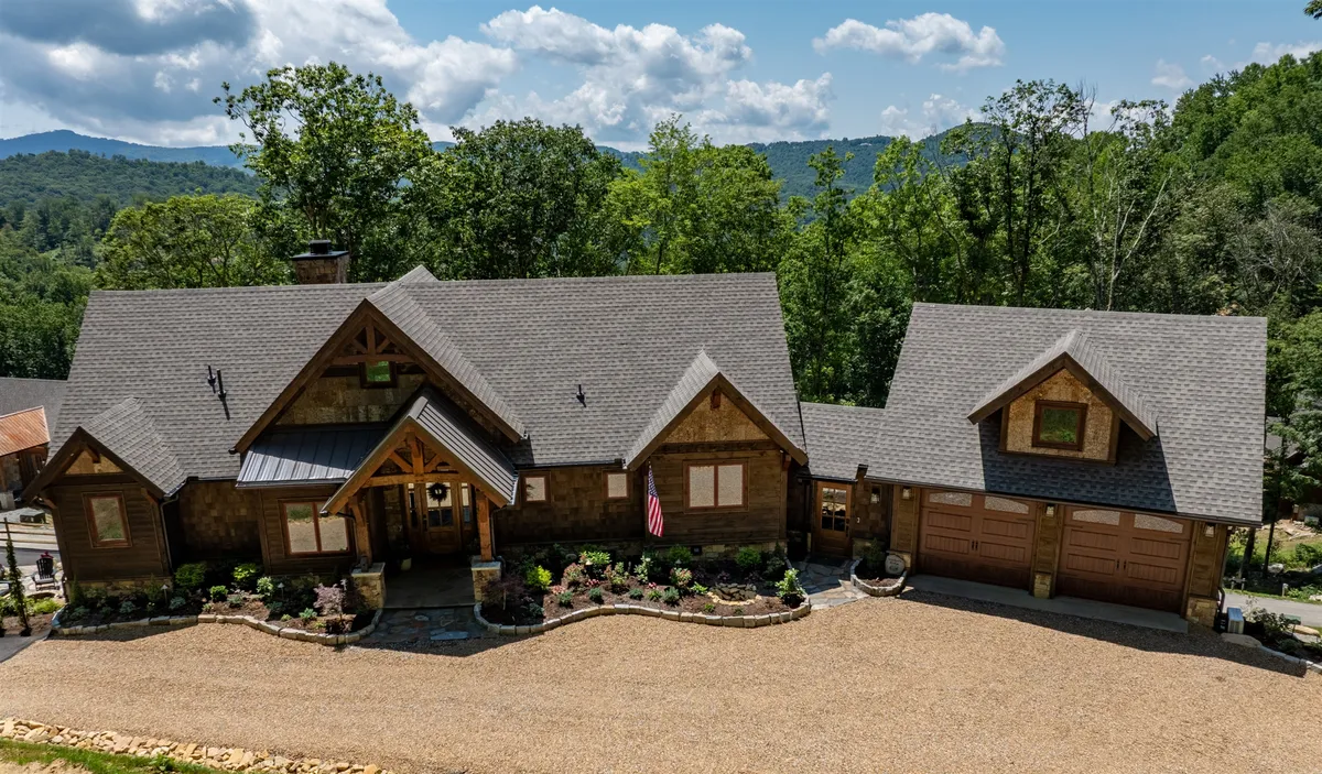 The image showcases a stunning mountain home crafted with warm wooden architecture, nestled into a lush, landscaped yard with panoramic mountain views behind. The inviting front porch hints at spaces perfect for family relaxation, morning coffee, or sunset gatherings, all set against the backdrop of the iconic North Carolina High Country scenery.