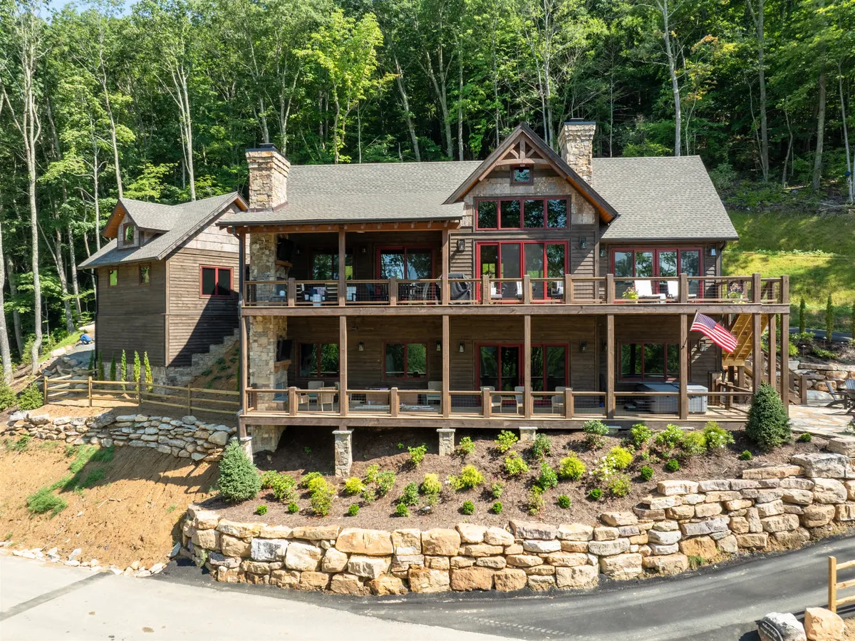 A large front porch with stone accent walls, red window frames, and surrounding greenery creates an inviting transition between indoor comfort and outdoor adventure. The expansive porch is perfect for winding down after a day on the trails, enjoying sunset views, or morning coffee amid nature.