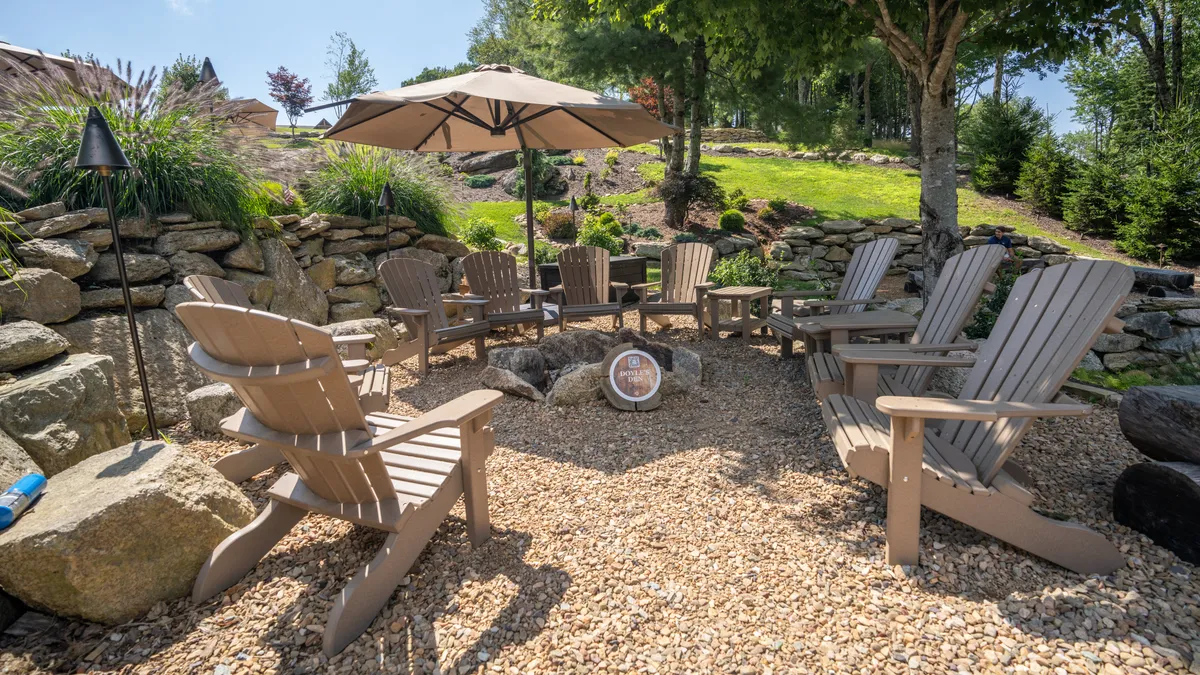 A cozy outdoor seating area featuring Adirondack chairs, a large umbrella, a crackling fire pit, and unique rock formations. The scene is set against a backdrop of trees, suggesting a tranquil setting for gathering, storytelling, and soaking in mountain views.