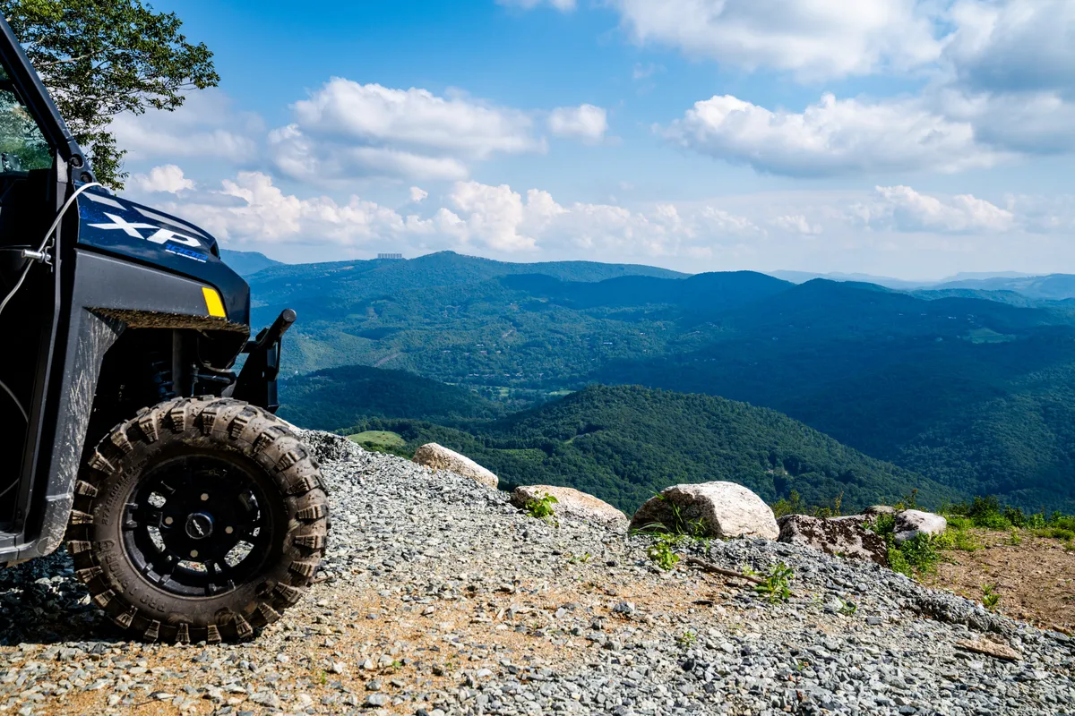 An off-road vehicle is pictured set against a dramatic mountain range and cloudy sky, suggesting the thrill of UTV rides and off-trail exploration. The open terrain and distant peaks evoke a sense of wild adventure and access to rugged trails.