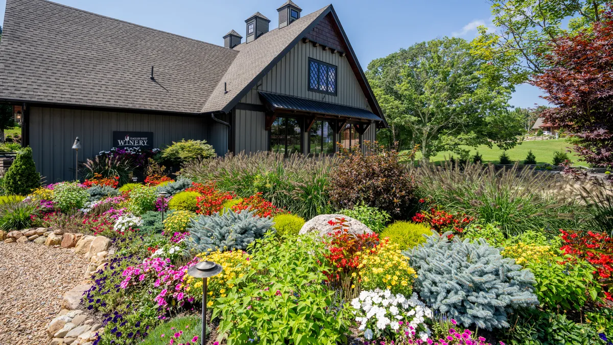 This image shows a vibrant exterior scene with lush flower beds, architectural details of the building, and surrounding greenery, suggesting a tranquil and inviting setting for outdoor gatherings and wine tasting.