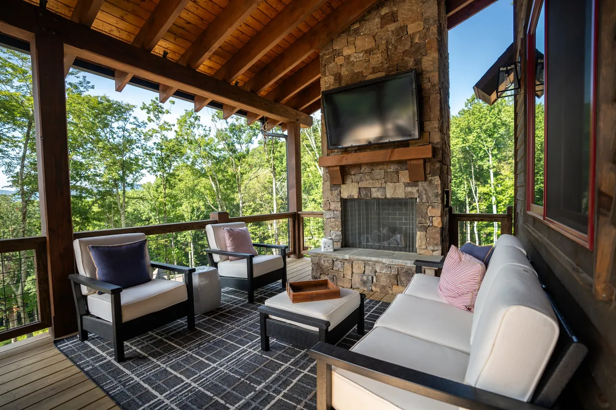 A covered outdoor deck with plush seating arranged around a stone fireplace, a television above the mantel, and natural greenery all around. The design invites relaxation and socializing with the comfort of indoor amenities and the beauty of the outdoors.