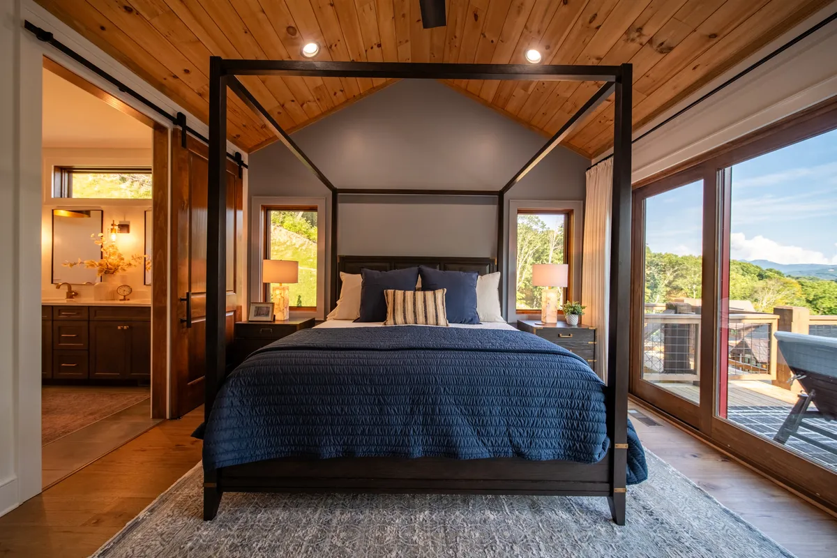 A serene bedroom featuring a king canopy bed positioned beside large windows that frame a mountain view. The space feels tranquil, with soft bedding and calming decor, and a doorway leads to a luxurious bathroom. Natural light fills the room, reinforcing the sense of privacy and comfort.