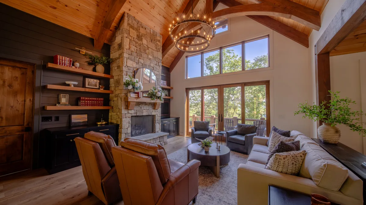 This interior shot centers on a spacious great room featuring a dramatic stone fireplace, large windows that draw in natural light, a striking chandelier, and an inviting seating arrangement. The room exudes warmth and comfort, offering scenic mountain views beyond the windows.