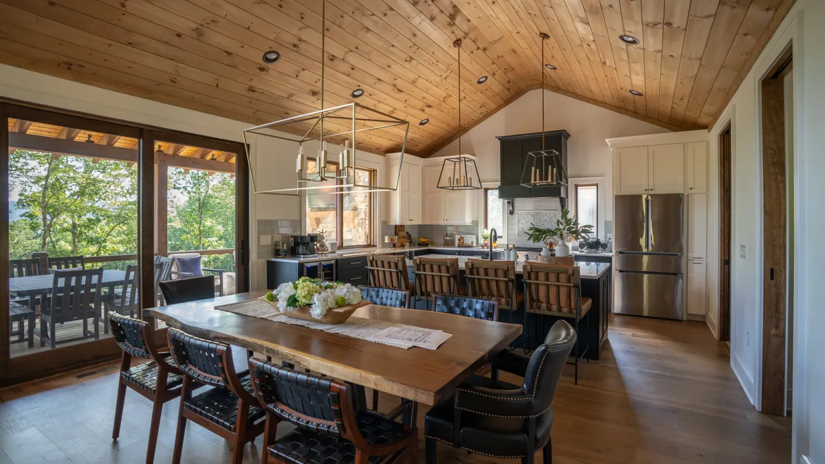 An open-concept interior showing a live-edge dining table, an island bar with seating, rich wooden ceiling details, and high-end kitchen cabinetry. The communal atmosphere is enhanced by the visible flow between kitchen, dining, and living areas.