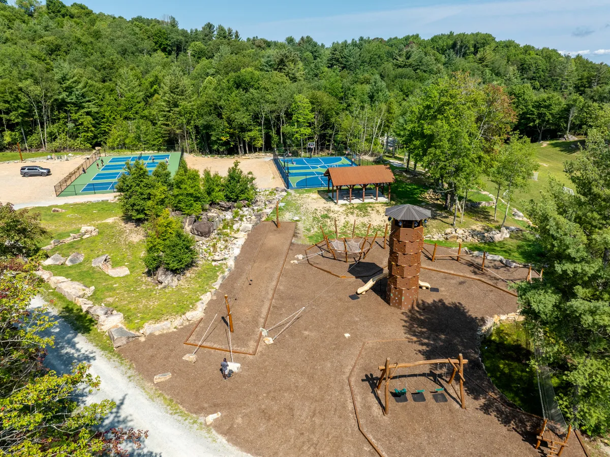 The image features a lively playground climbing structure, adjacent tennis courts, a pavilion, and children actively playing outdoors. Families can be seen gathering and enjoying the open landscape, with the natural surroundings providing a vibrant backdrop.